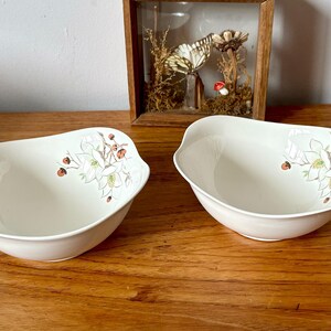 May include: Two white ceramic bowls with a floral design. The bowls have a white background with a floral pattern in shades of green, brown, and pink. The bowls are slightly oval shaped and have a rounded lip.