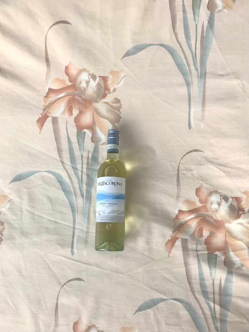 May include: A bottle of Mezzacorona Pinot Grigio wine on a white background with a floral pattern. The bottle is clear glass with a white label and a gold screw top.
