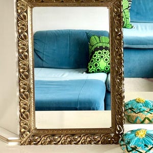 May include: An ornate gold rectangular mirror with detailed scrollwork. The mirror reflects a blue sofa and patterned cushions. Decorative gold-toned baskets with blue and yellow accents are in the foreground. A classic home decor piece.
