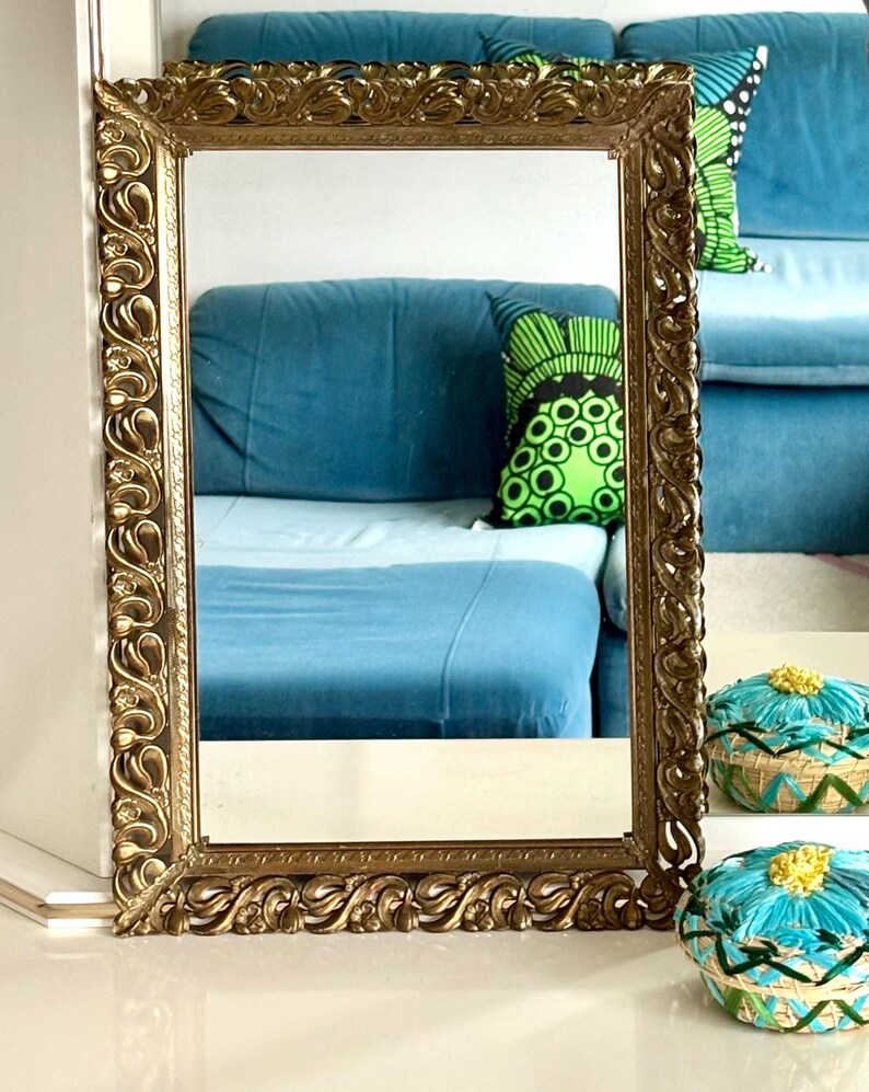May include: An ornate gold rectangular mirror with detailed scrollwork. The mirror reflects a blue sofa and patterned cushions. Decorative gold-toned baskets with blue and yellow accents are in the foreground. A classic home decor piece.