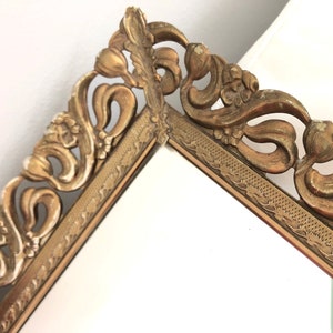 May include: Close-up of a vintage gold mirror frame. The frame features elaborate floral detailing and a decorative border. The mirror is set against a white background, highlighting the intricate design of the frame.