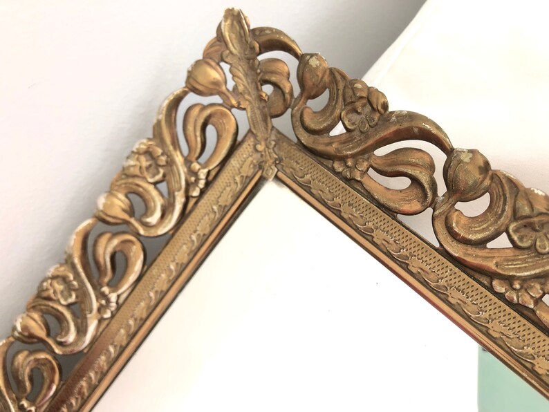 May include: Close-up of a vintage gold mirror frame. The frame features elaborate floral detailing and a decorative border. The mirror is set against a white background, highlighting the intricate design of the frame.