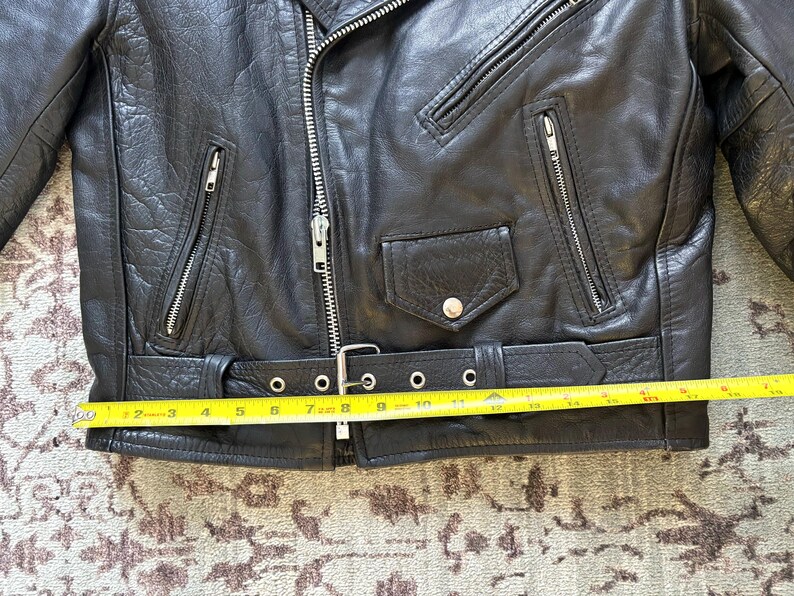 May include: Black leather motorcycle jacket with a zipper closure, two pockets, and a belt with a buckle. The jacket is measured with a yellow tape measure.