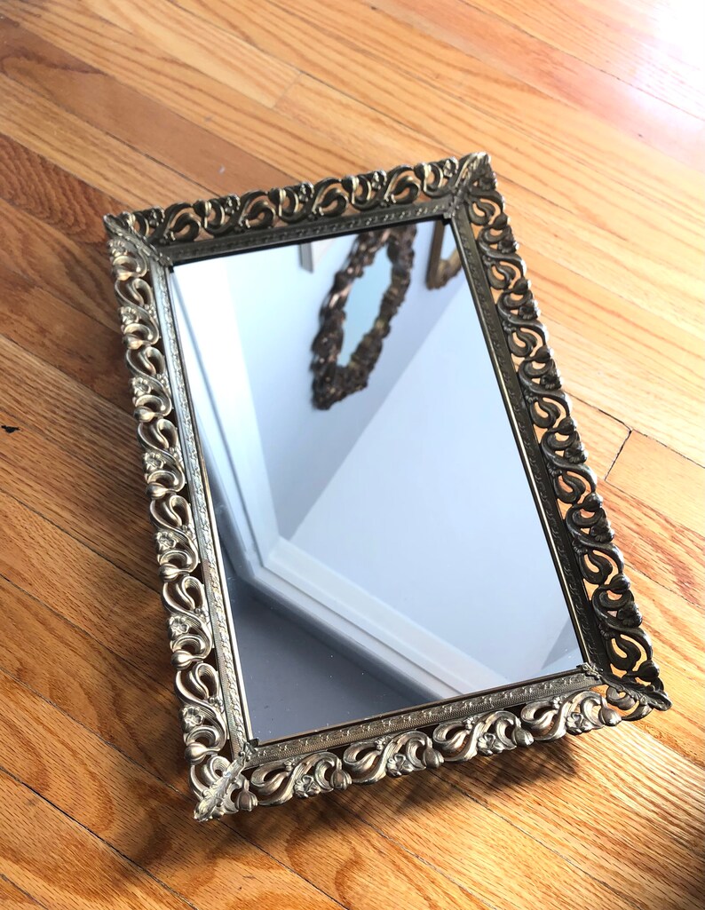 May include: A rectangular gold-toned mirror with an ornate, scrolling floral design frame. The mirror reflects a partial view of a room. The frame has a vintage aesthetic, adding a touch of elegance to any space.