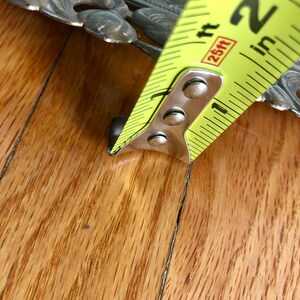 May include: A yellow and black tape measure showing a measurement of approximately 5 cm. The tape measure is positioned next to a decorative metal object with floral designs. The background is a wooden surface.