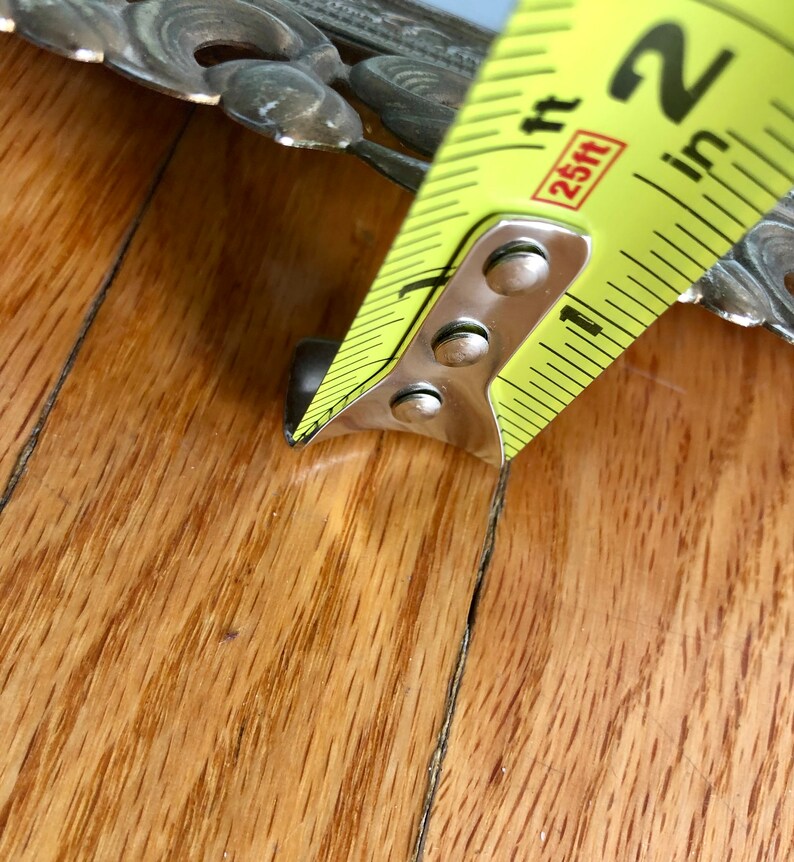 May include: A yellow and black tape measure showing a measurement of approximately 5 cm. The tape measure is positioned next to a decorative metal object with floral designs. The background is a wooden surface.