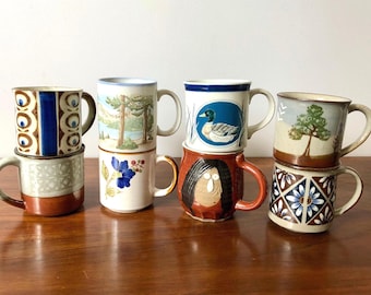 May include: A collection of seven ceramic mugs arranged in two stacks. The mugs feature various designs including a blue duck, a tree, a blue flower, a face, and geometric patterns in shades of brown, blue, white, and green.