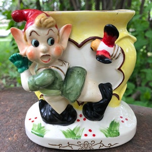 May include: A vintage ceramic planter featuring a whimsical elf with a red hat and green clothing, holding a bird in his hand. The planter is yellow with a white background and red and green accents.