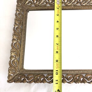 May include: An antique rectangular vanity mirror with an ornate, gold-coloured metal frame. The mirror measures approximately 30 cm in length. The frame features a detailed, scrolling floral design.