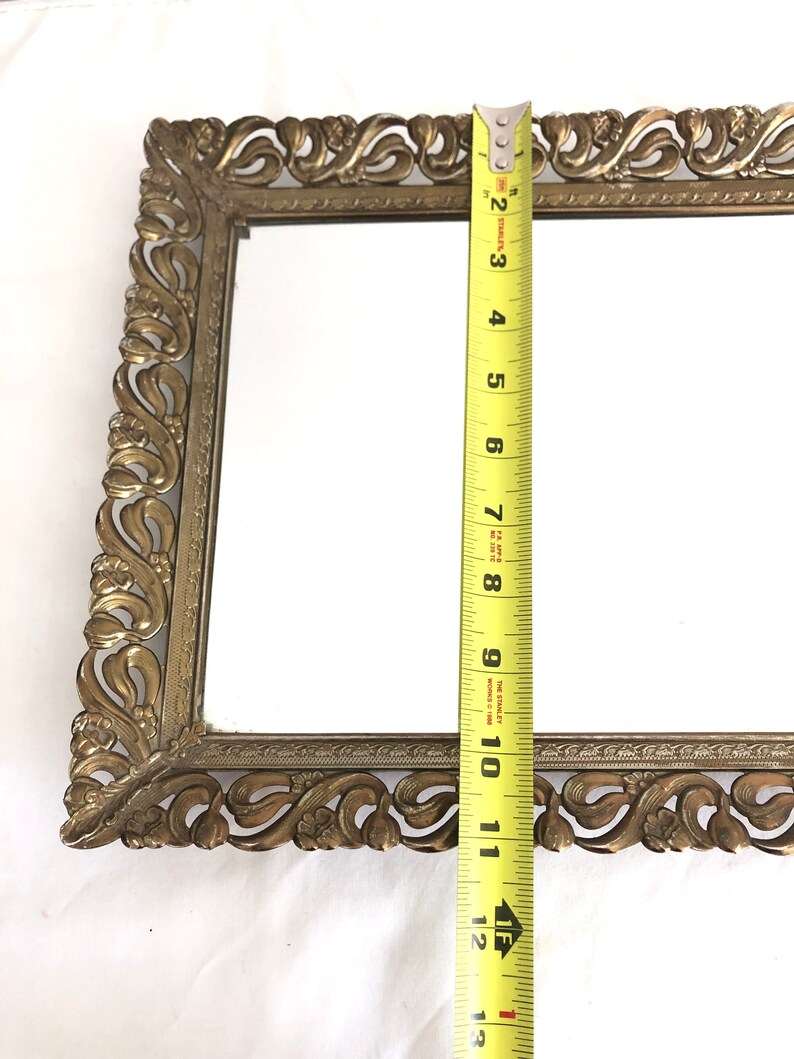 May include: An antique rectangular vanity mirror with an ornate, gold-coloured metal frame. The mirror measures approximately 30 cm in length. The frame features a detailed, scrolling floral design.