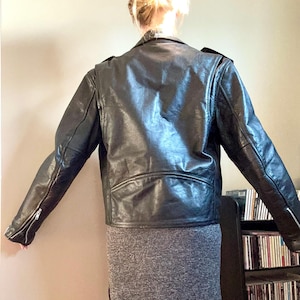 May include: Black leather motorcycle jacket with a zipper closure and a stand-up collar. The jacket has a slightly fitted silhouette and is shown on a model wearing a gray skirt.
