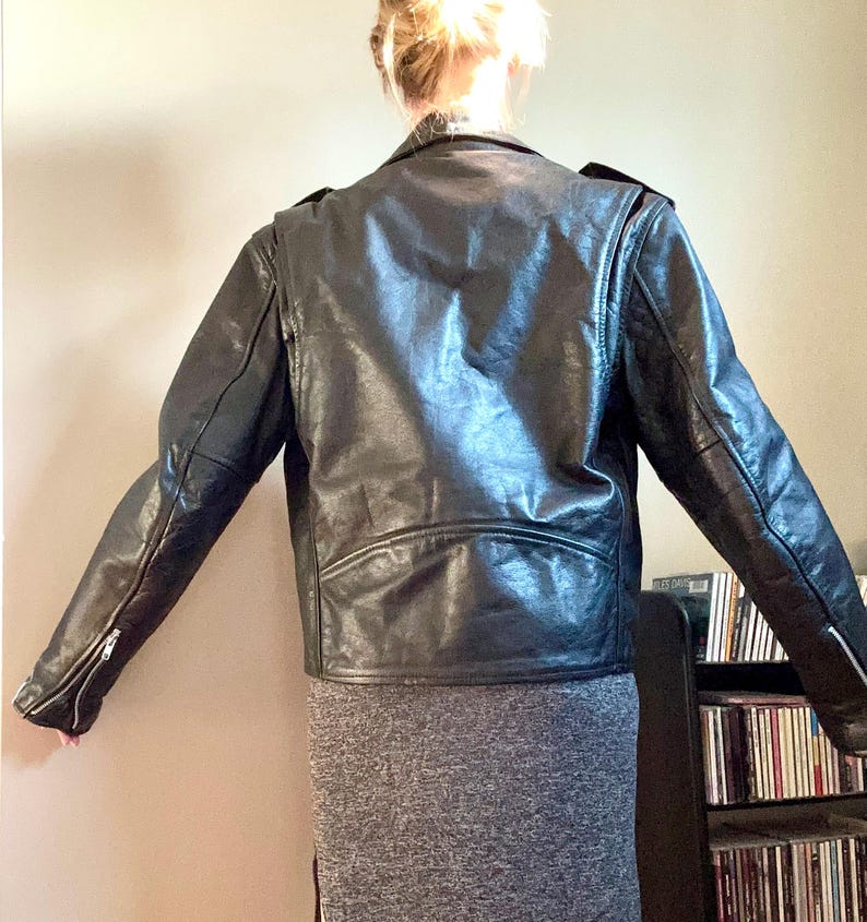 May include: Black leather motorcycle jacket with a zipper closure and a stand-up collar. The jacket has a slightly fitted silhouette and is shown on a model wearing a gray skirt.