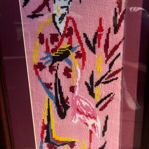 May include: A framed needlepoint embroidery depicting a woman in a colourful kimono standing next to a pink flamingo on a pink background.