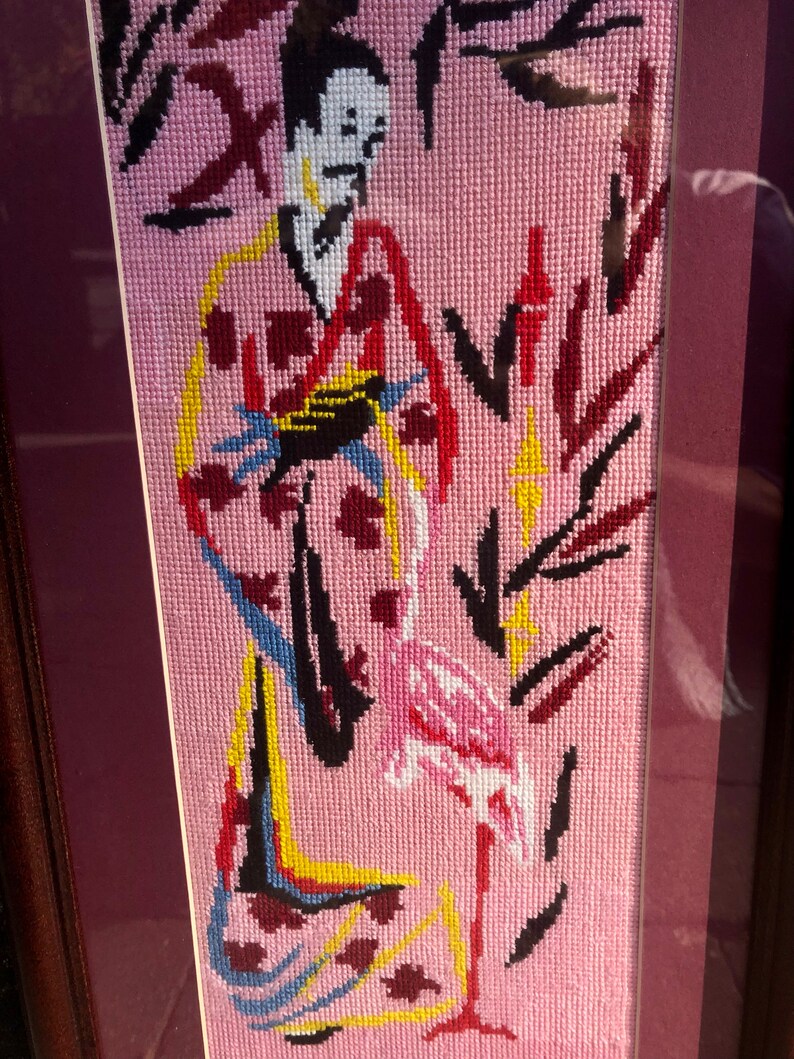 May include: A framed needlepoint embroidery depicting a woman in a colourful kimono standing next to a pink flamingo on a pink background.