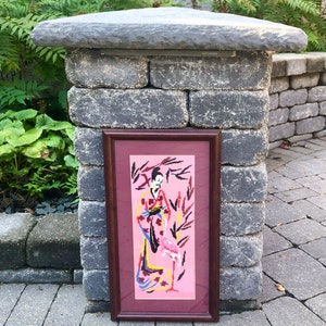 May include: A framed needlepoint artwork depicting a woman in a pink kimono standing with a pink flamingo in a pink and green landscape.