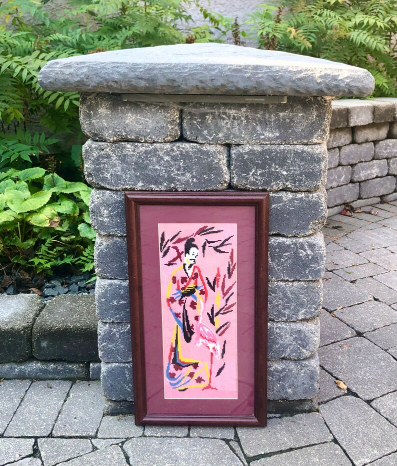 May include: A framed needlepoint artwork depicting a woman in a pink kimono standing with a pink flamingo in a pink and green landscape.