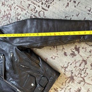 May include: Black leather motorcycle jacket with a zipper closure and multiple pockets. The jacket has a quilted lining on the sleeves and a yellow measuring tape is placed across the sleeve to show the length.
