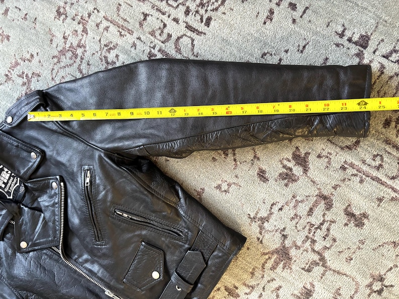 May include: Black leather motorcycle jacket with a zipper closure and multiple pockets. The jacket has a quilted lining on the sleeves and a yellow measuring tape is placed across the sleeve to show the length.