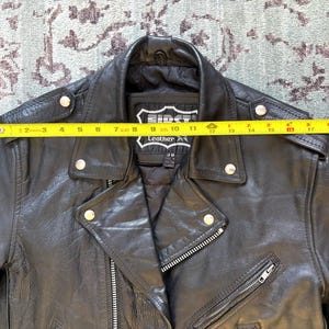 May include: Black leather motorcycle jacket with a zipper closure and multiple pockets. The jacket has a label that reads "First Leather Apparel".