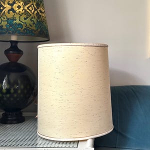 May include: A cylindrical lamp shade with a cream-coloured fabric exterior featuring a subtle speckled pattern. The shade has a simple, straight-sided design with finished edges at the top and bottom, providing a neutral and clean appearance.