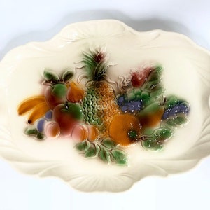 May include: A white ceramic serving tray featuring a raised design of fruit, including a pineapple, bananas, grapes, and apples. The fruit is painted in shades of green, yellow, orange, red, and purple.