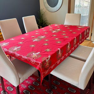 May include: A red rectangular tablecloth with a festive design featuring gold and white accents, including bells and decorative elements. The tablecloth is draped over a wooden table, surrounded by beige chairs, and set on a red patterned rug.