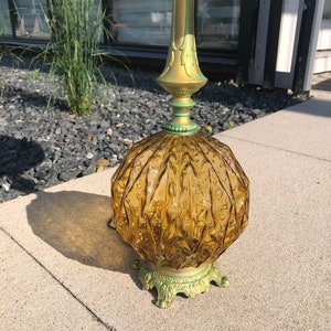 Pu&ograve; includere: Una lampada da tavolo con una base in vetro color ambra, di forma rotonda e con una texture a coste. La lampada presenta uno stelo metallico color oro e una base decorativa e ornata con accenti di patina verde, che sostiene un paralume bianco.