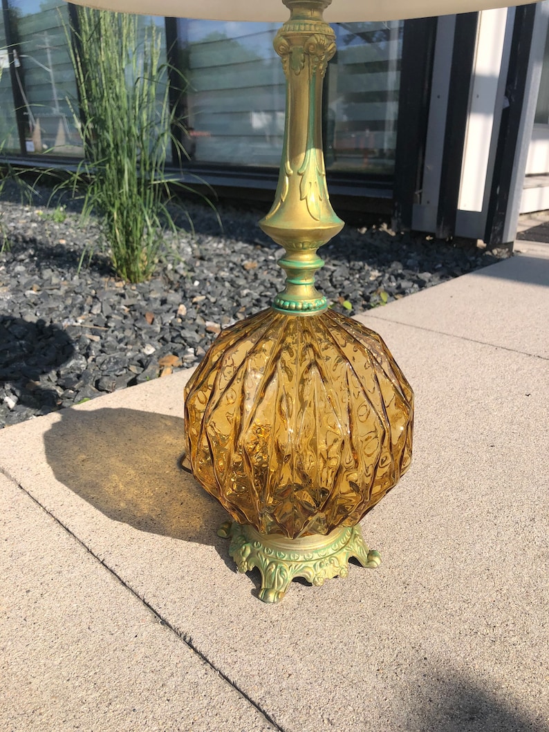 Pu&ograve; includere: Una lampada da tavolo con una base in vetro color ambra, di forma rotonda e con una texture a coste. La lampada presenta uno stelo metallico color oro e una base decorativa e ornata con accenti di patina verde, che sostiene un paralume bianco.