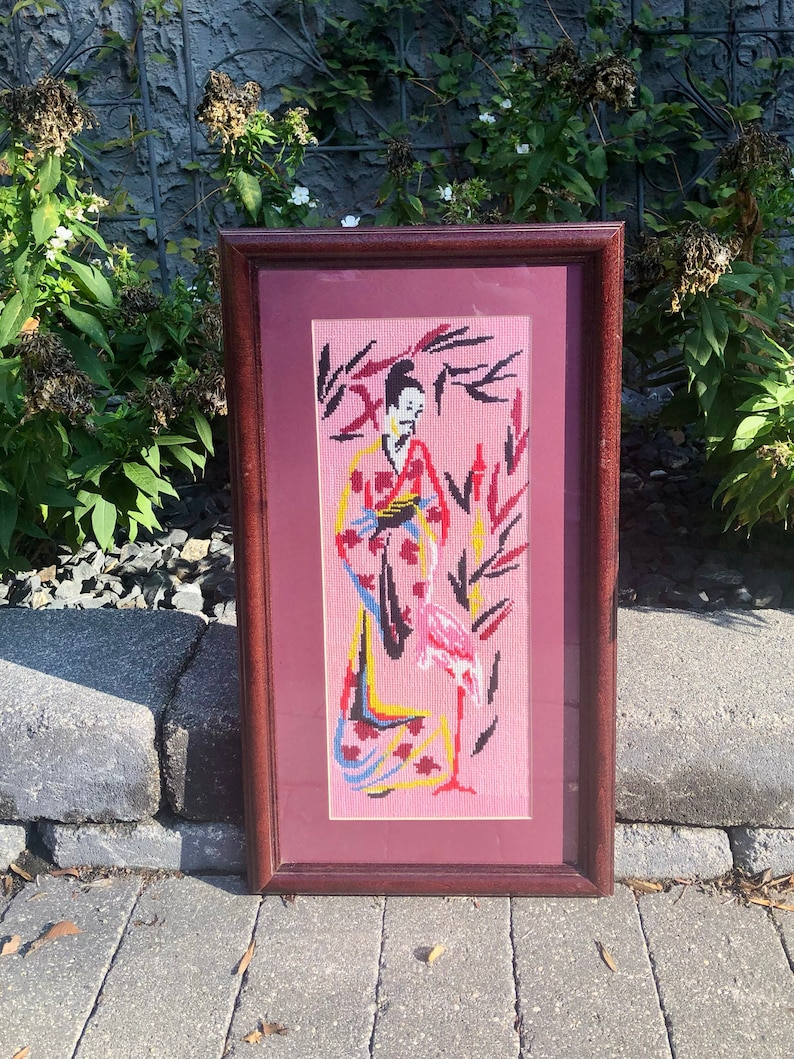 May include: Framed needlepoint artwork depicting a woman in a colourful kimono standing with a pink flamingo on a pink background.