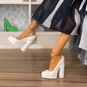 Può includere: Scarpe bianche con tacco alto e tacco a blocco. Le scarpe sono indossate da una bambola, con un vestito a righe bianche e nere. Una scarpa con tacco alto verde è visibile sullo sfondo. Le scarpe sono su una superficie di legno.