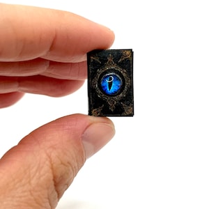 May include: Miniature black book with a blue eye design on the cover. The eye is detailed with a white highlight. The book has gold-colored filigree around the eye and on the corners. The book is held between two fingers.