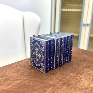 Pode incluir: Uma pilha de livros azuis em miniatura com detalhes dourados e desenhos intrincados. Os livros estão dispostos em uma fila sobre uma superfície de madeira.