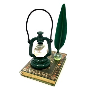 May include: A miniature green lantern with a black handle, a green feather quill, and a small book with gold-colored edges. The lantern and quill are positioned on top of the book, creating a vintage aesthetic.