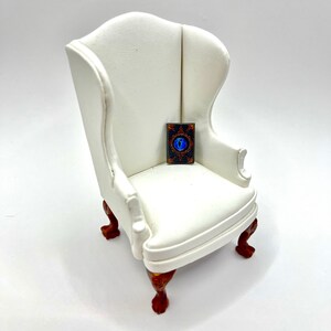 May include: A miniature white wingback chair with dark wood legs and a small rectangular object on the seat. The object has a dark background with a blue eye design and gold accents.