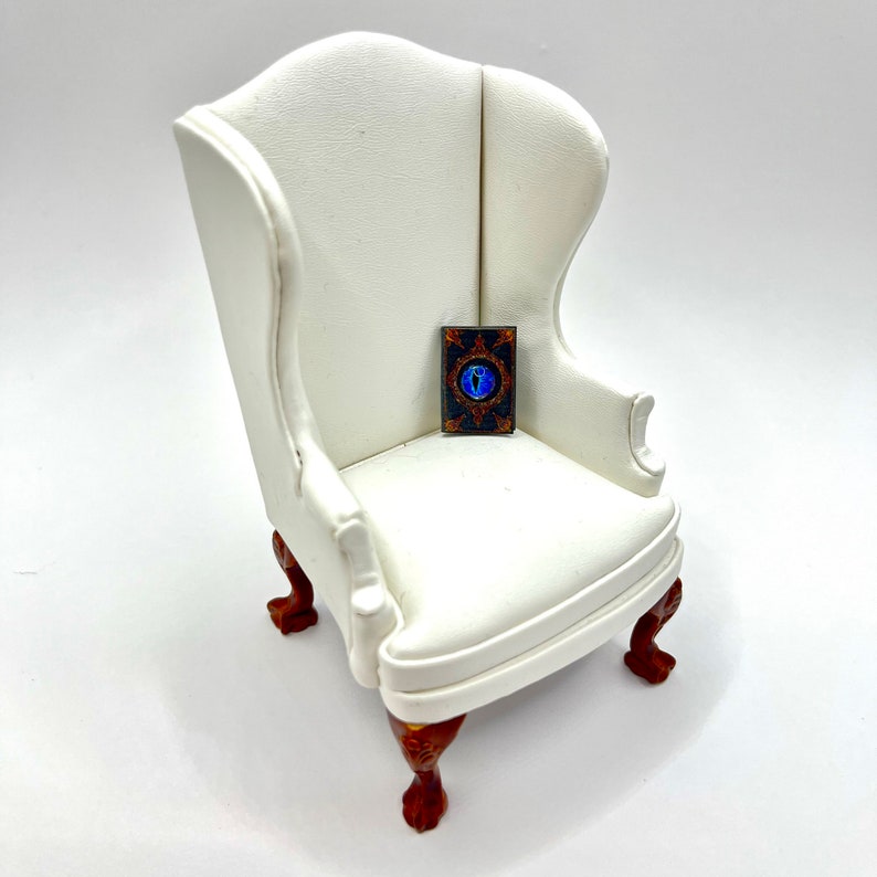 May include: A miniature white wingback chair with dark wood legs and a small rectangular object on the seat. The object has a dark background with a blue eye design and gold accents.