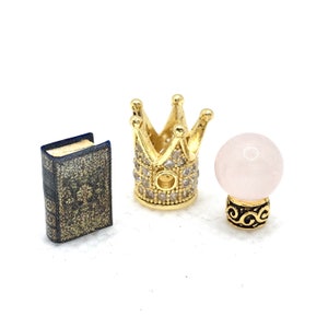 Può includere: Una corona dorata con strass trasparenti, una piccola sfera di cristallo rosa con base dorata e un piccolo libro nero con dettagli dorati.