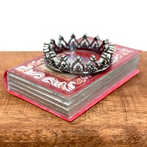 May include: A black metal crown ring with a delicate design, resting on top of a red book with ornate details.