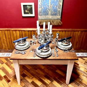 Könnte beinhalten: Ein Miniatur-Silberleuchter mit sechs Kerzenständern und einer silbernen Basis steht auf einem Holztisch mit einer weißen Tischdecke. Der Tisch ist mit zwei Gedecken gedeckt, jedes mit einem silbernen Teller, einer silbernen Schüssel, einer silbernen Gabel, einem silbernen Messer, einem silbernen Löffel und einem silbernen Weinglas. Jedes Gedeck hat auch eine blaue Serviette, die aufgerollt und auf den Teller gelegt wird.