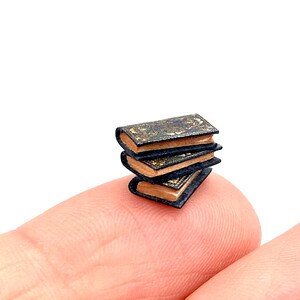May include: A stack of three miniature books with black covers and gold accents. The books are sitting on a finger.