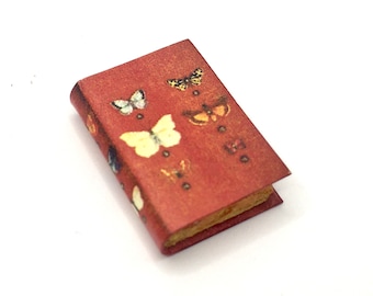 Può includere: Un piccolo libro rosso con un design a farfalla. La copertina presenta una varietà di farfalle in diversi colori e motivi.
