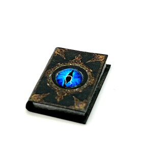 May include: A miniature black book with a blue eye design on the cover. The eye is set within a gold-colored frame with ornate detailing. The book's edges have a gold trim.