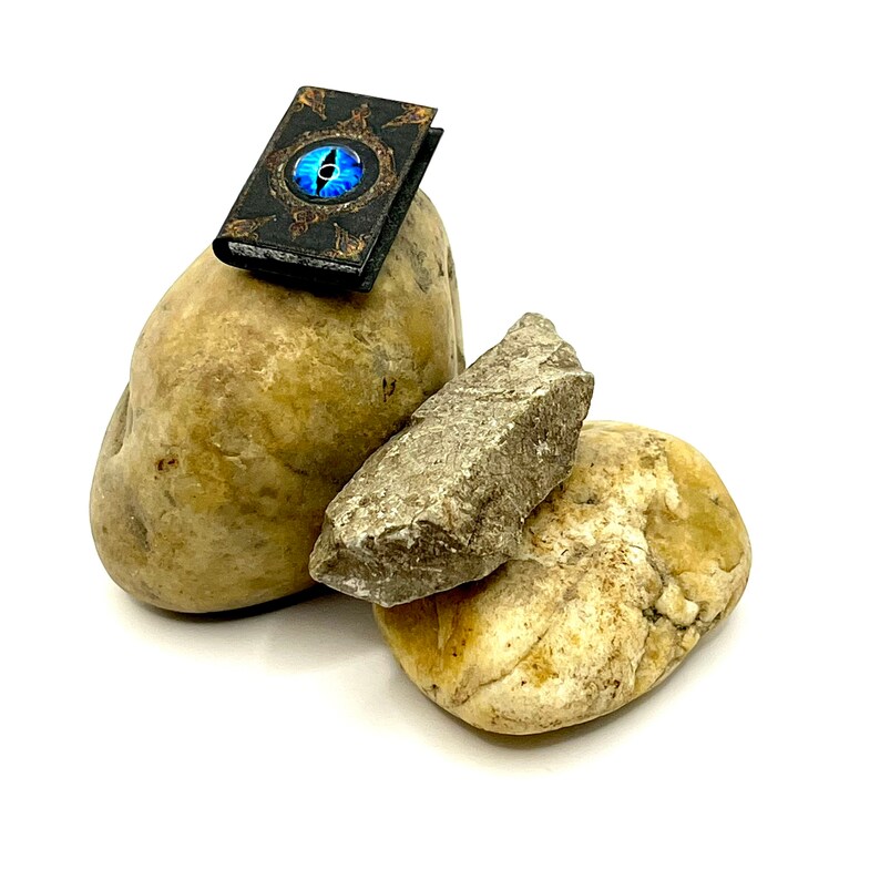 May include: A miniature book with a blue eye design on the cover rests on a light brown stone. The book has a black cover with gold accents. Two other stones, one light brown and one gray, are also in the image.