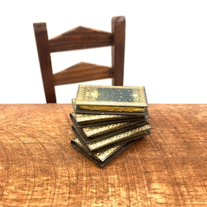 May include: A stack of small, antique books with gold-leaf covers and black spines sits on a wooden table. The books are arranged in a neat pile, with the top book facing the viewer.