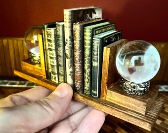 Könnte beinhalten: Ein Miniatur-Holzregal mit einer Reihe von antiken Büchern und zwei Glas-Schneekugeln. Das Regal ist braun lackiert und hat ein dekoratives Design. Die Bücher haben aufwendige Illustrationen und Titel wie "Winter's Tale" und "The Painter's Elements".