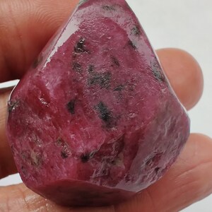 May include: A large, rough, pink ruby gemstone with black inclusions. The ruby is faceted and has a glossy finish.