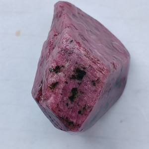 Puede incluir: Un gran cristal de rubí rosa en bruto con inclusiones negras. El cristal tiene una forma angular y puntiaguda.