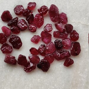 May include: A collection of small, rough, red gemstones. The gemstones are scattered on a white surface.