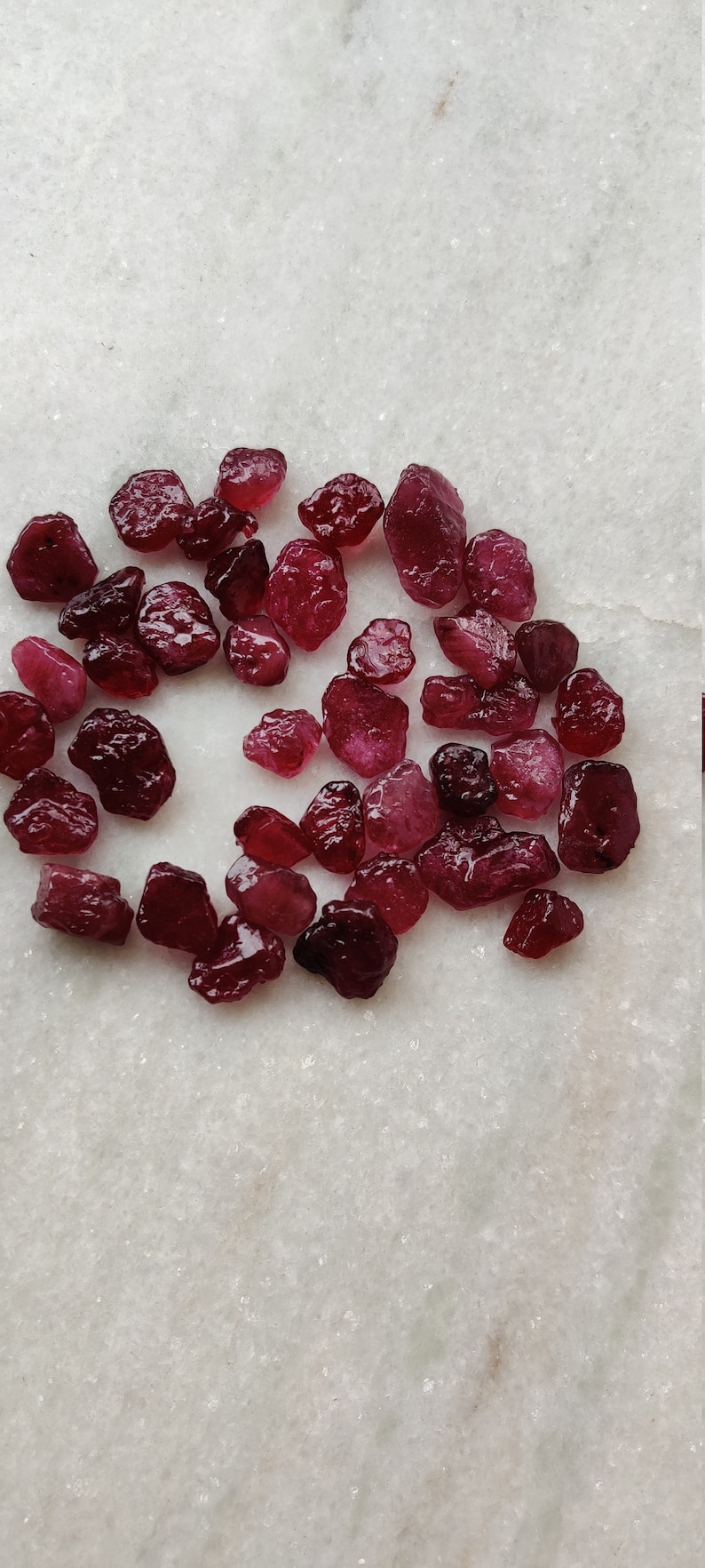 May include: A collection of small, rough, red gemstones. The gemstones are scattered on a white surface.