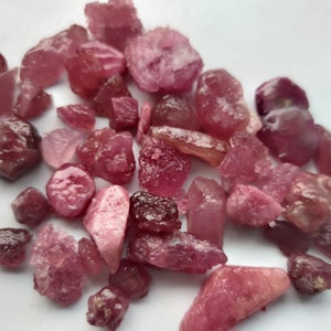 May include: A collection of raw, uncut pink tourmaline gemstones. The stones vary in size and shape, displaying a range of pink hues from light to deep rose. The rough texture and natural forms of the crystals are visible.