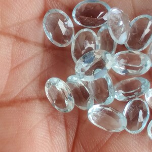 May include: A scattering of oval-cut, pale blue gemstones. The faceted stones are displayed on a palm, highlighting their clarity and colour. These gemstones are likely for jewellery making.
